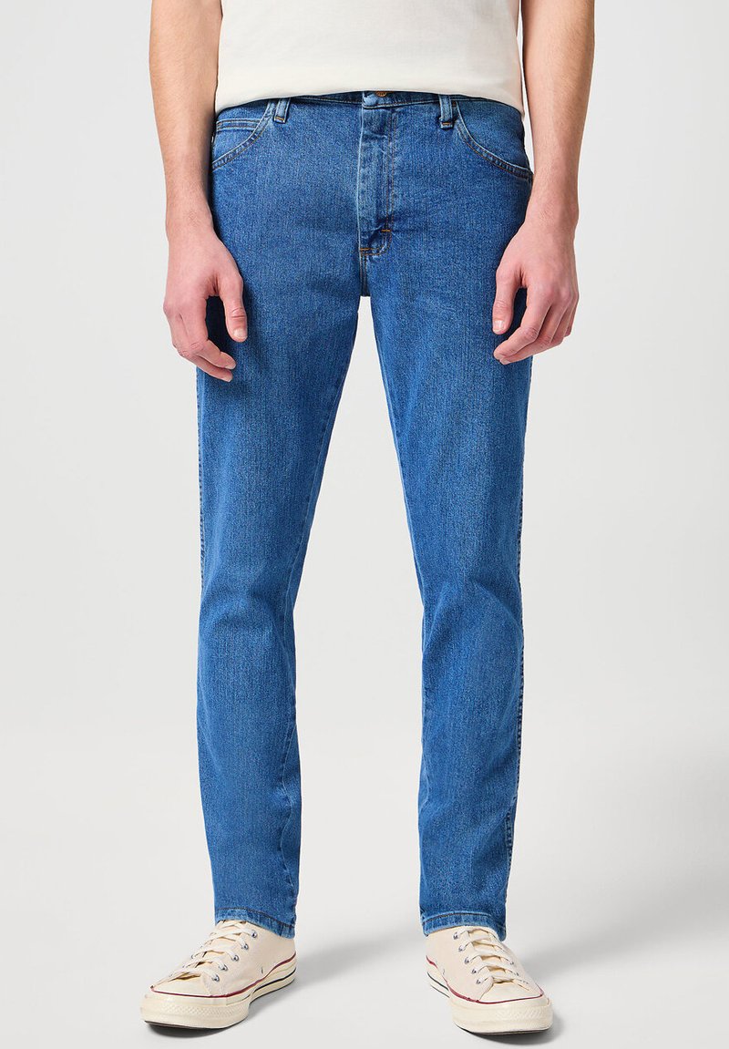 Niebieskie dżinsy o prostym kroju, z delikatnym wyblaknięciem, pięcioma kieszeniami i metalowymi nitami, w zestawieniu z kremowymi sneakersami.