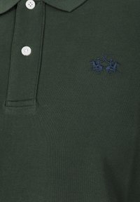 Tmavě zelené polo tričko s texturovaným povrchem, na němž je vyšitá námořnicky modrá značka se dvěma jezdeckými postavami a u límce jsou tři bílé knoflíky.