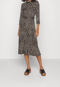 Schwarz-beiges Kleid mit Leopardenmuster, dreiviertellangen Ärmeln, tailliertem Gürtel und fließendem Saum, kombiniert mit schwarzen Clogs mit Holzabsätzen.