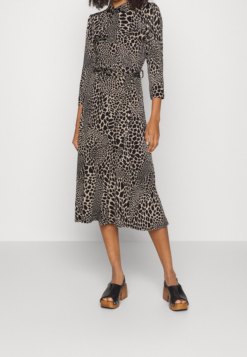 Schwarz-beiges Kleid mit Leopardenmuster, dreiviertellangen Ärmeln, tailliertem Gürtel und fließendem Saum, kombiniert mit schwarzen Clogs mit Holzabsätzen.