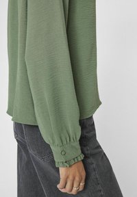 Grüne Textur-Bluse mit langen Ärmeln, gerafften Manschetten und lockerer Passform, kombiniert mit dunkelschwarzen Jeans. Minimalistisches Design und subtile Details.
