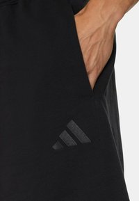 Bermudas deportivas negras de tela suave, con un bolsillo lateral y un sutil logo de tres rayas grises en la parte inferior izquierda.