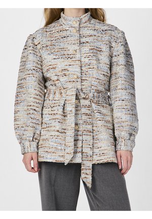 Kobieta nosząca teksturowaną, wielokolorową marynarkę z tweedu ze złotymi guzikami i pasem wiązanym w talii, zestawioną z szarymi spodniami.