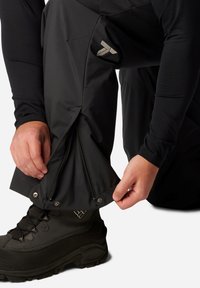 Wasserdichte schwarze Hosen mit Reißverschlüssen an den Knöcheln und silbernen Akzenten. Eine Hand stellt den Reißverschluss ein, während ein schwarzer Stiefel darunter sichtbar ist.