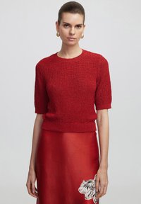 Czerwony sweter z dzianiny z krótkimi rękawami i ściągaczem, w połączeniu z czerwoną satynową spódnicą z kwiatowym wzorem. Gładka tekstura i dopasowany krój.