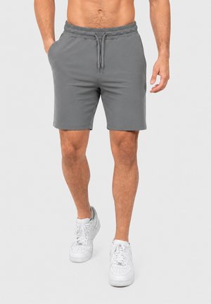 Homme portant un short gris à cordon et des baskets blanches, debout avec une main dans la poche devant un fond uni.