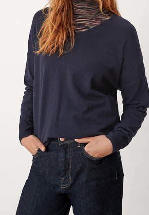 Pull à manches longues marine avec un col en V, fabriqué en tissu lisse. Associé à un jean en denim foncé avec des accents cuivrés et une taille cintrée.