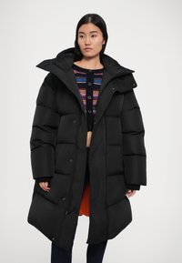 Abrigo acolchado negro con capucha, forma oversized, textura acolchada y cremallera frontal. Presenta un forro naranja en contraste y puños acanalados.