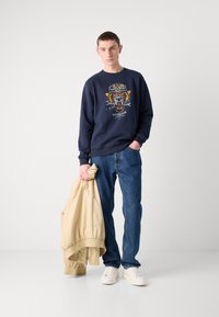 Navy sweatshirt with tiger graphic, blue jeans, and beige jacket, held in left hand. White sneakers complete the outfit.
