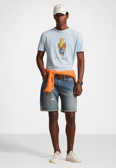 T-shirt de algodão azul claro com impressão gráfica, suéter de malha laranja amarrado na cintura, calções de ganga desgastados e sapatilhas off-white.