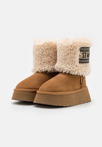 Stivali in suede marrone con un morbido risvolto in pelliccia shearling color crema. Suola spessa e stratificata con una finitura testurizzata. Etichetta laterale con un design a righe.