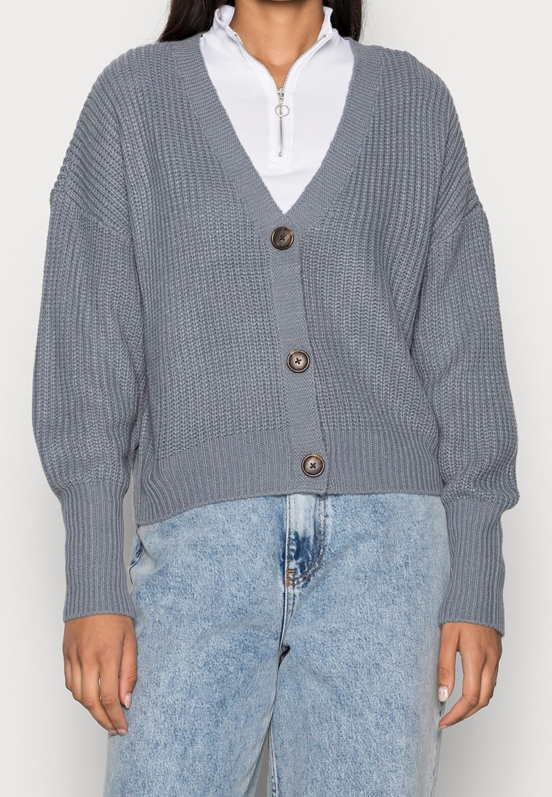 Szary dziergany kardigan z dekoltem w serek, w luźnym kroju, z trzema brązowymi guzikami. Projekt zawiera prążkowane rękawy i teksturowane wykończenie.