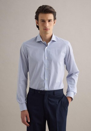 Giovane uomo con camicia a righe bianco e blu infilata in pantaloni blu scuro, in piedi con una mano in tasca davanti a uno sfondo semplice.