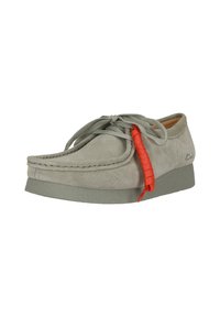 Chaussure en suede gris clair avec un bout arrondi, des détails de coutures surélevés et un accent de lacet rouge contrastant. Caractérisée par une semelle en caoutchouc gris texturé.