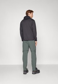 Svart hettejakke, lett stoff, sammen med grønne bukser med glidelåser og lommer, og svarte teksturerte joggesko.