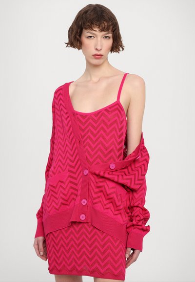 Completo lavorato a maglia di colore rosa composto da un vestito senza maniche e un cardigan oversize con motivo a zigzag. Il cardigan presenta grandi bottoni e polsini a costine.
