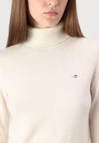 Jasno beżowy sweter golfowy wykonany z miękkiego materiału. Posiada małe haftowane logo na piersi oraz żebrowany detal przy szyi.