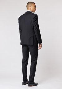 Czarny garnitur o dopasowanej sylwetce, z klapami w kształcie notcha i pojedynczym pęknięciem z tyłu. Stylizowany z czarnymi eleganckimi butami i spodniami.
