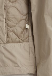Helle beige Pufferjacke mit gestepptem Design, ausgestattet mit einem kleinen Logoetikett und glattem Material. Genähte Nähte entlang des Randes.