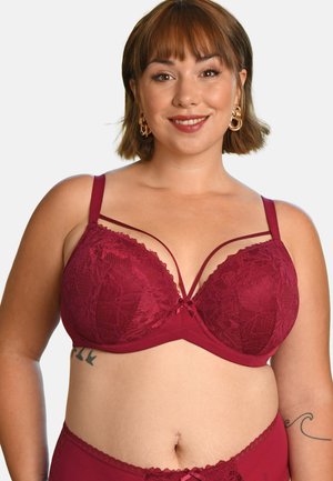 Smiling woman with short brown hair wearing burgundy lace bra and matching bottoms, gold hoop earrings, and visible tattoos on torso and arm.