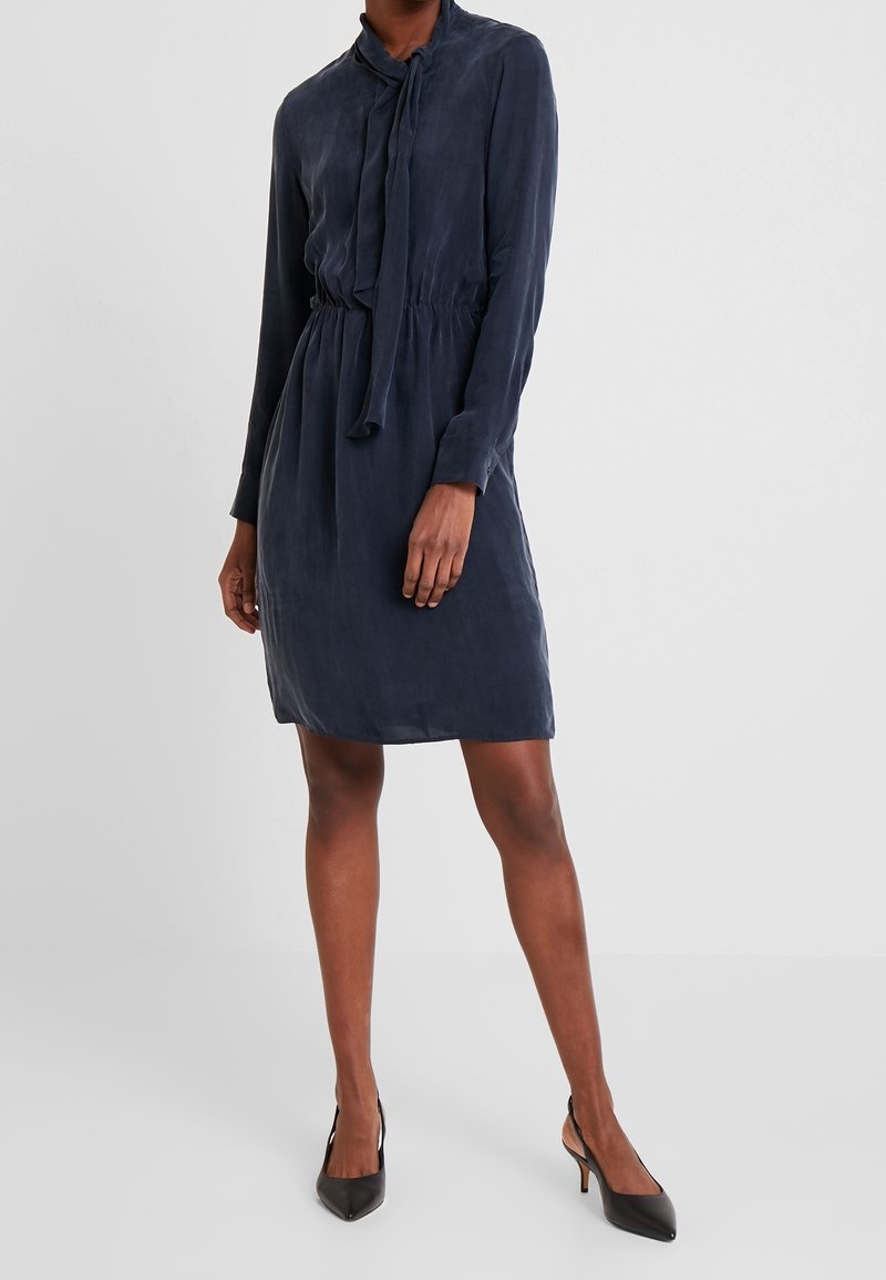 Abito blu navy con maniche lunghe, dettaglio al collo con fiocco, vita elastica e texture liscia. Abbinato a scarpe con tacco nere.