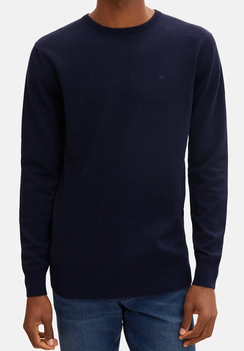 Granatowy sweter, okrągły dekolt, długie rękawy, gładka tekstura, ściągacze przy rękawach i u dołu, subtelny detal z logo na klatce piersiowej, w połączeniu z dżinsami.