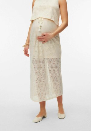 Femme enceinte portant une robe en maille beige sans manches avec des pampilles, tenant son ventre, debout dans des chaussures crème à talons bas.