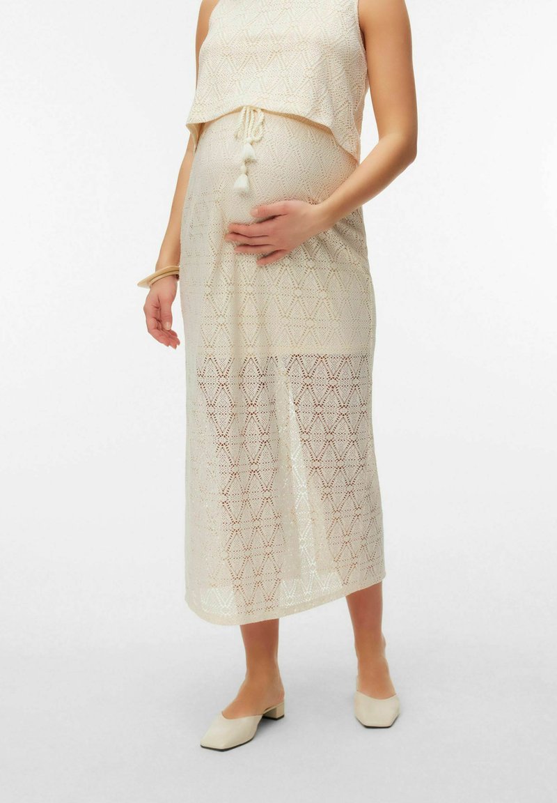 Femme enceinte portant une robe en maille beige sans manches avec des pampilles, tenant son ventre, debout dans des chaussures crème à talons bas.
