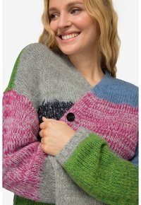 Monivärinen neulecardigan, jossa on harmaan, vaaleanpunaisen, sinisen, vihreän ja mustan värisiä paneeleja. Kankaan pinta on teksturoitu, ja se sulkeutuu edestä napilla.