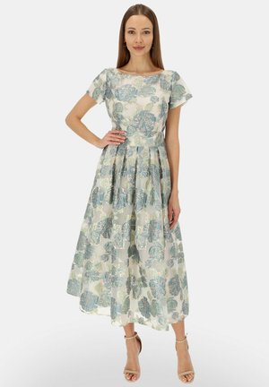 Kobieta w krótkim, midi sukience w kwiatowy wzór w kolorach niebieskim i jasnej zieleni, stojąca z jedną ręką na biodrze, w beżowych szpilkach.