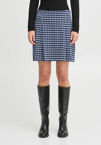 Jupe à carreaux bleus et blancs avec des plis, en tissu, associée à des bottes en cuir noires jusqu'aux genoux.