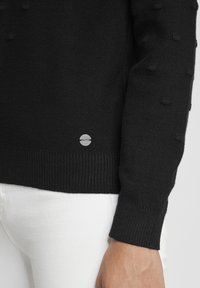 Pulover negru tricotat cu texturi bombate pe mâneci, cu manșete din elastic și un logo mic circular la tiv. Asortat cu blugi albi.