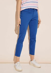 Blue, slim-fit trousers with a smooth texture, front pockets, and two accent buttons, paired with white sneakers and a striped top.