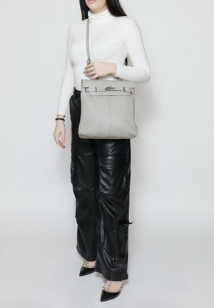 Donna che indossa un dolcevita bianco e pantaloni di pelle neri, tiene una borsa crossbody grigia e indossa décolleté nere con punta a punta e cinturini alla caviglia.