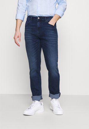 Homme portant un jean bleu foncé retroussé aux chevilles, des baskets blanches Adidas et une chemise à manches longues bleu clair, une main dans la poche.
