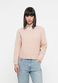 Lys rosa genser, rund hals, myk tekstur, vide ermer, båret over en collared stripet skjorte, kombinert med lyseblå jeans.