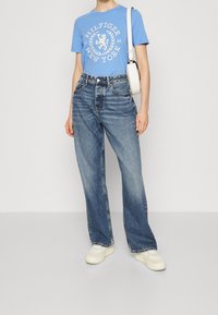 En person som bär en ljusblå grafisk t-shirt, högmidjade lösa blå jeans, vita plattformssneakers och bär en vit axelväska.