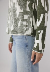 Grün-weißer gemusterter Langarmshirt, kombiniert mit hellblauen Jeans. Verfügt über einen runden Ausschnitt und dezente Markenlogos an der Seite.