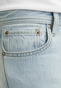 Vaqueros de mezclilla azul claro con un acabado texturizado, que incluyen un pequeño bolsillo frontal, remaches de bronce y costuras amarillas en contraste.