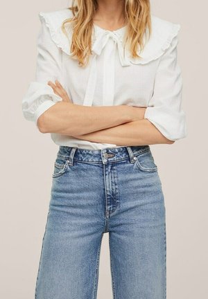 Femme portant un chemisier blanc à col à volants et nœud noué, ainsi qu'un jean bleu taille haute, les mains croisées sur la taille.