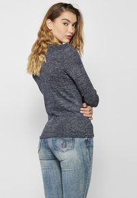 Mėlynasis ilgarankovis marškinėlis su tekstūruotu mezgimo dizainu, derinamas su šviesiai mėlynomis džinsinėmis kelnėmis, turinčiomis nugarinius kišenes su siuvinėjimo elementais.