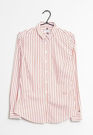 Biała koszula na długi rękaw z pionowymi pomarańczowymi paskami, małe haftowane "T.H." blisko dołu, wisząca na czarnym wieszaku na białym tle.