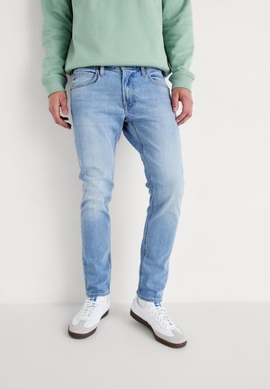 Homme portant un jean slim bleu clair, des baskets blanches et un sweat-shirt vert clair, debout devant un fond blanc uni.