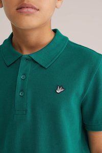 Zöld póló, texturált anyagból, gallérral és három gombbal, bal mellen egy kis hímzett madárlogóval.