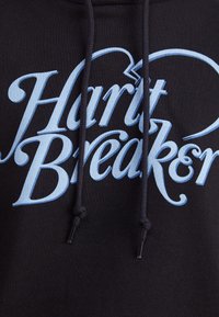 Svart hettegenser med "Heartbreaker" i lys blå kursiv tekst. Har snor med metalltupper og en teksturert stoff.