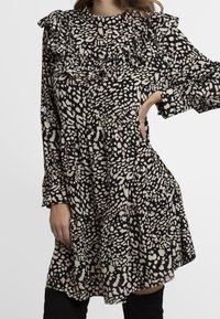 Robe à motifs noir et beige avec un col volanté et des manches longues, présentant une jupe à volants et une texture de tissu léger.
