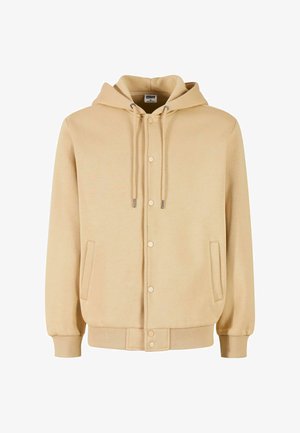 Chaqueta con capucha beige, con cremallera completa y cierre de botón, cordones ajustables, puños acanalados y un bolsillo frontal. Textura suave.