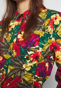 Chemise à boutons à motif floral présentant des couleurs vives comme le rouge, le jaune et le vert, avec de grands motifs floraux superposés et un col classique.