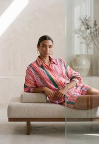 Žena s tmavými vlasmi v farebnom pruhovanom tričku a šortkách, sediaca uvoľnene na béžovom modernom ležadle v minimalistickej miestnosti.