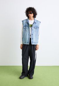 Gilet en denim avec de grandes emmanchures, couleur bleu clair, et boutons en métal, superposé sur une chemise blanche à manches longues et un pantalon noir. Lunettes de soleil noires incluses.